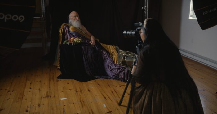Un homme avec une barbe est installé sur une chaise et costumé d'une autre époque pendant qu'il est photographié par une jeune femme à l'avant-plan.